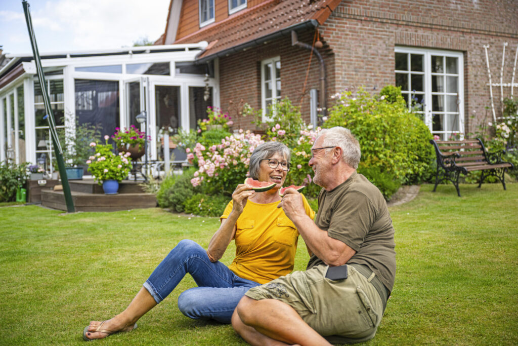ILLUSTRATION - Ein aelteres Paerchen sitzt am 18.07.2023 in einem Garten in Uelsby auf dem Rasen und isst Melone (gestellte Szene)
